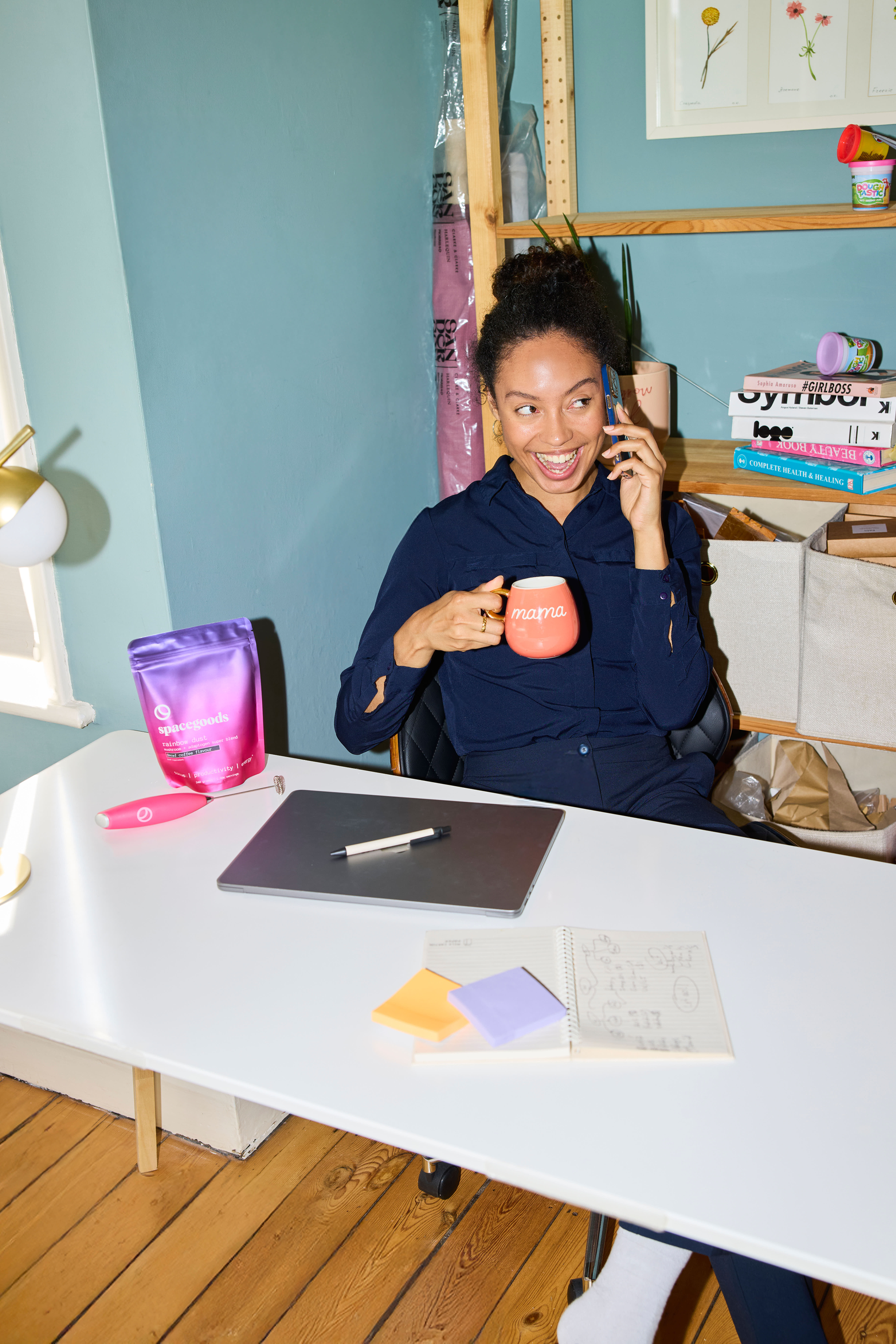 Woman sitting at a desk holding a mug and talking on a phone, with a bag of Spacegoods Rainbow Dust and stationery items around.